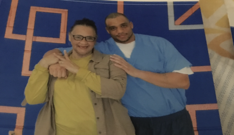 A man in prison-issued clothing stands with his arm around his mother.